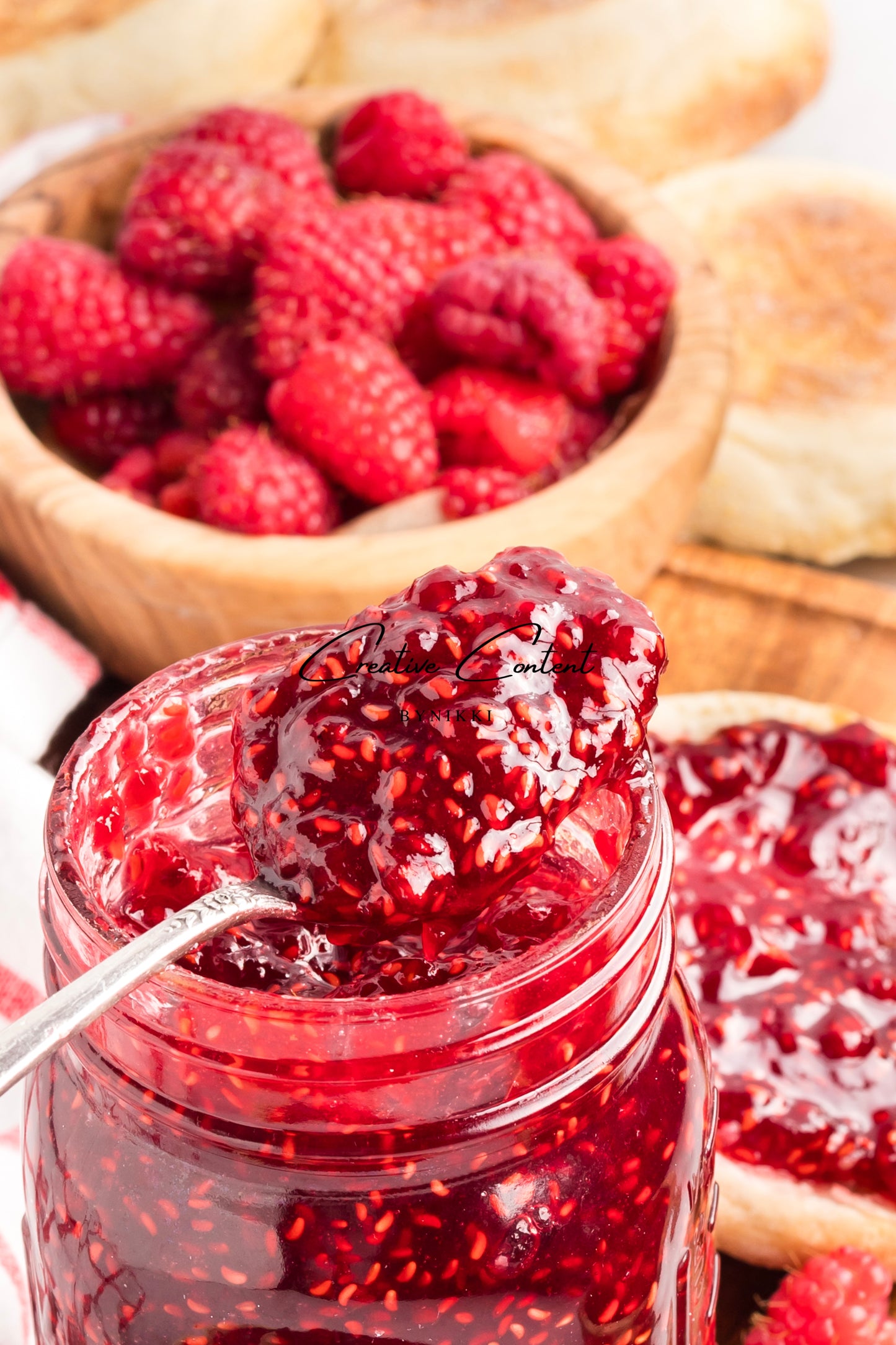 Jar of raspberry jam with a spoon, surrounded by fresh raspberries and bread.