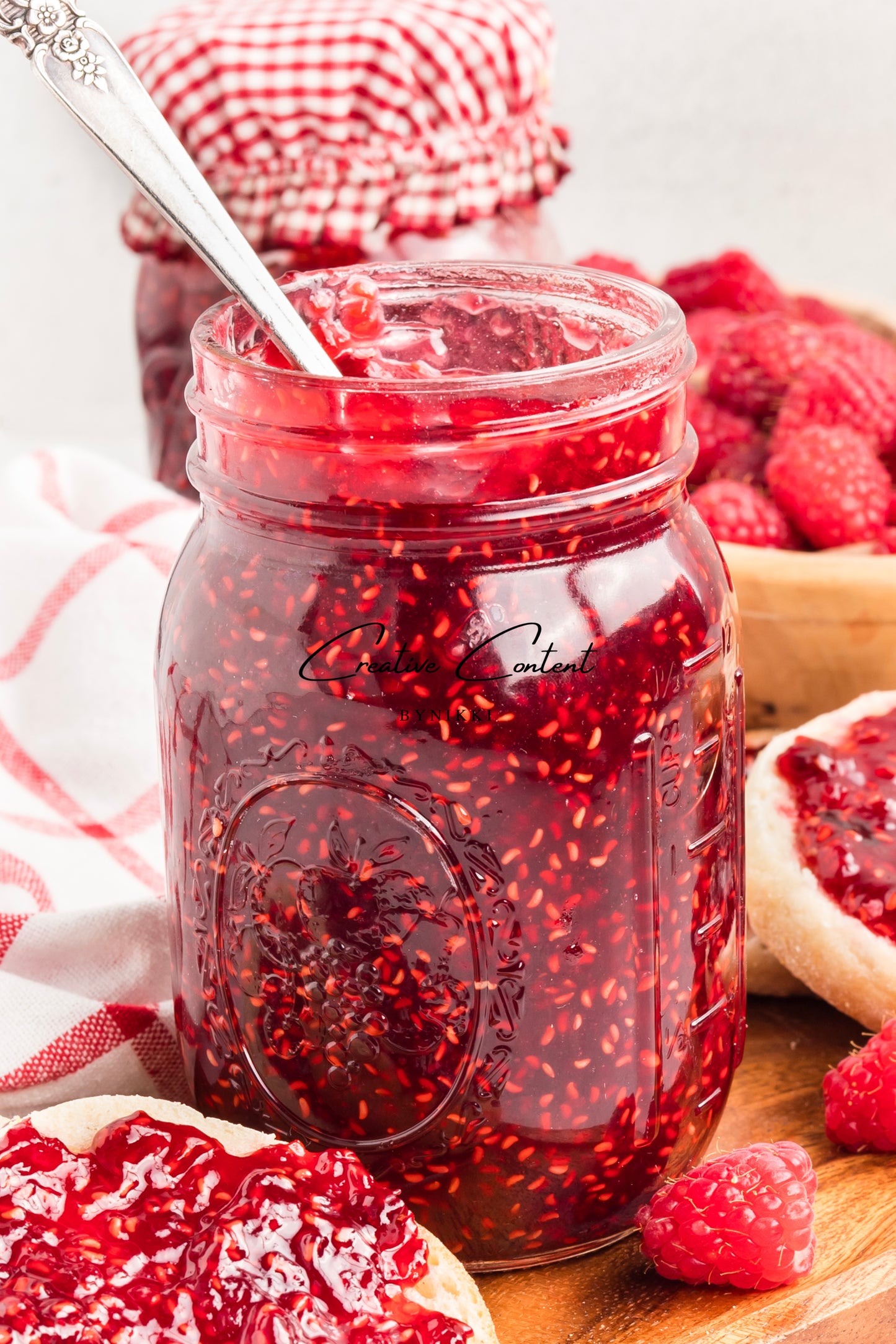 Jar of raspberry jam with a spoon, surrounded by raspberries and a checkered cloth.