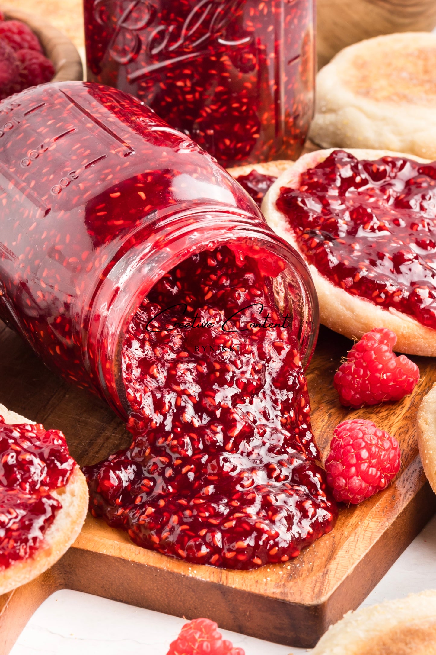 Jar of raspberry jam with a spreadable layer on a wooden board, surrounded by raspberries and bread.