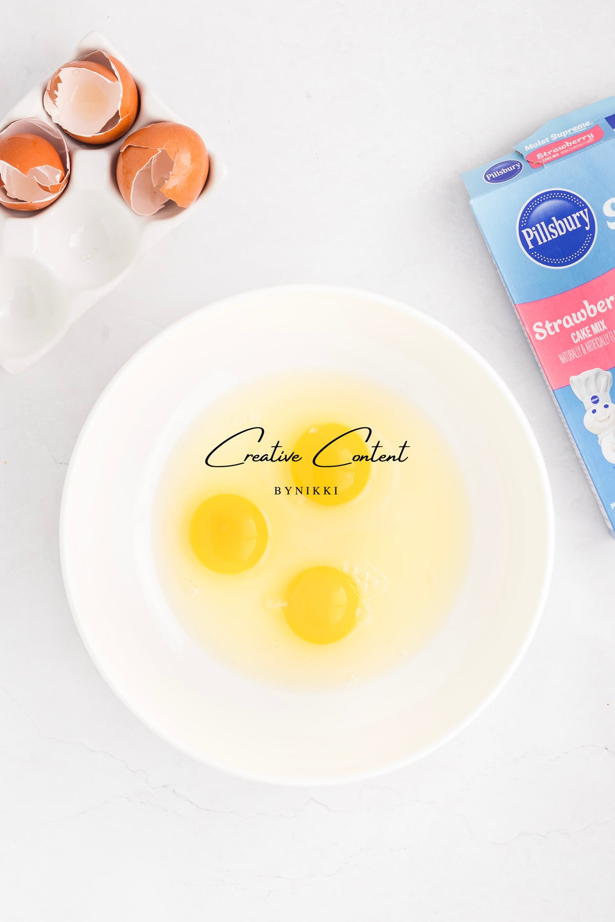 A white bowl containing three cracked eggs, positioned next to a carton of Strawberry Crunch Cake - Exclusive and a tray of empty eggshells.