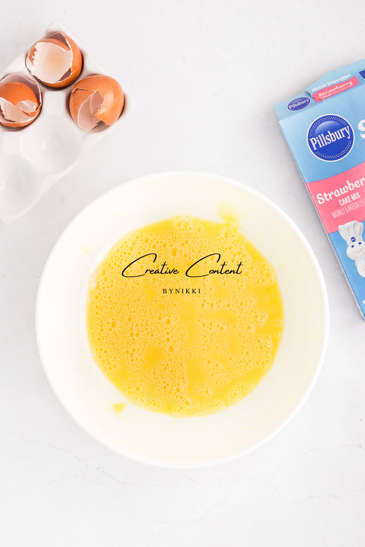 A white bowl containing whisked eggs for a Strawberry Crunch Cake - Exclusive, next to a carton of cake mix and broken eggshells.
