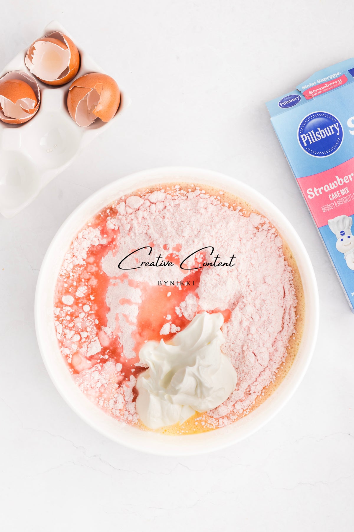 A mixing bowl contains ingredients for Strawberry Crunch Cake - Exclusive, including pink cake mix, liquid, and sour cream, with eggshells and the mix box nearby.