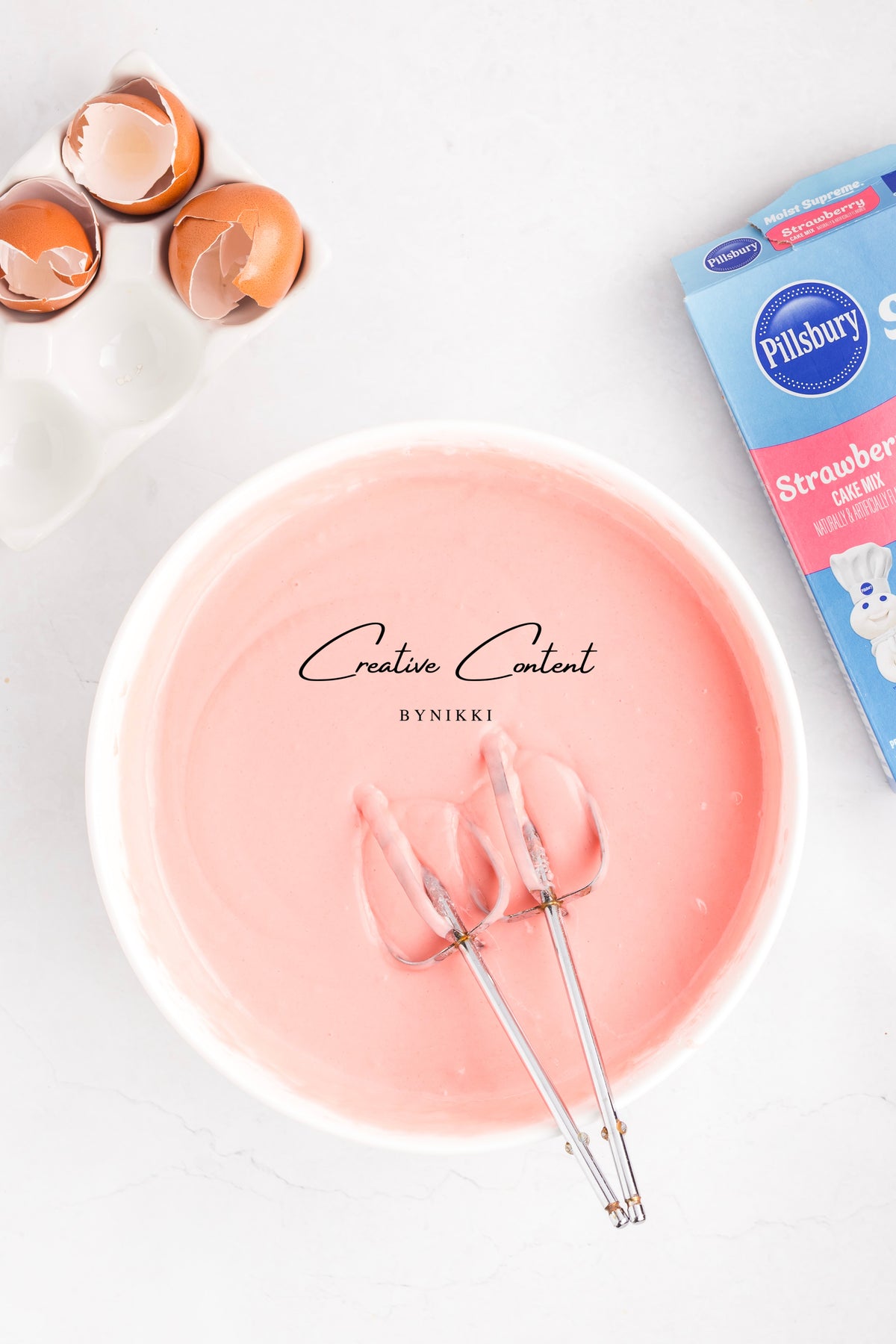 A white bowl filled with Strawberry Crunch Cake - Exclusive batter and two electric mixer beaters, next to a box of cake mix and empty eggshells.