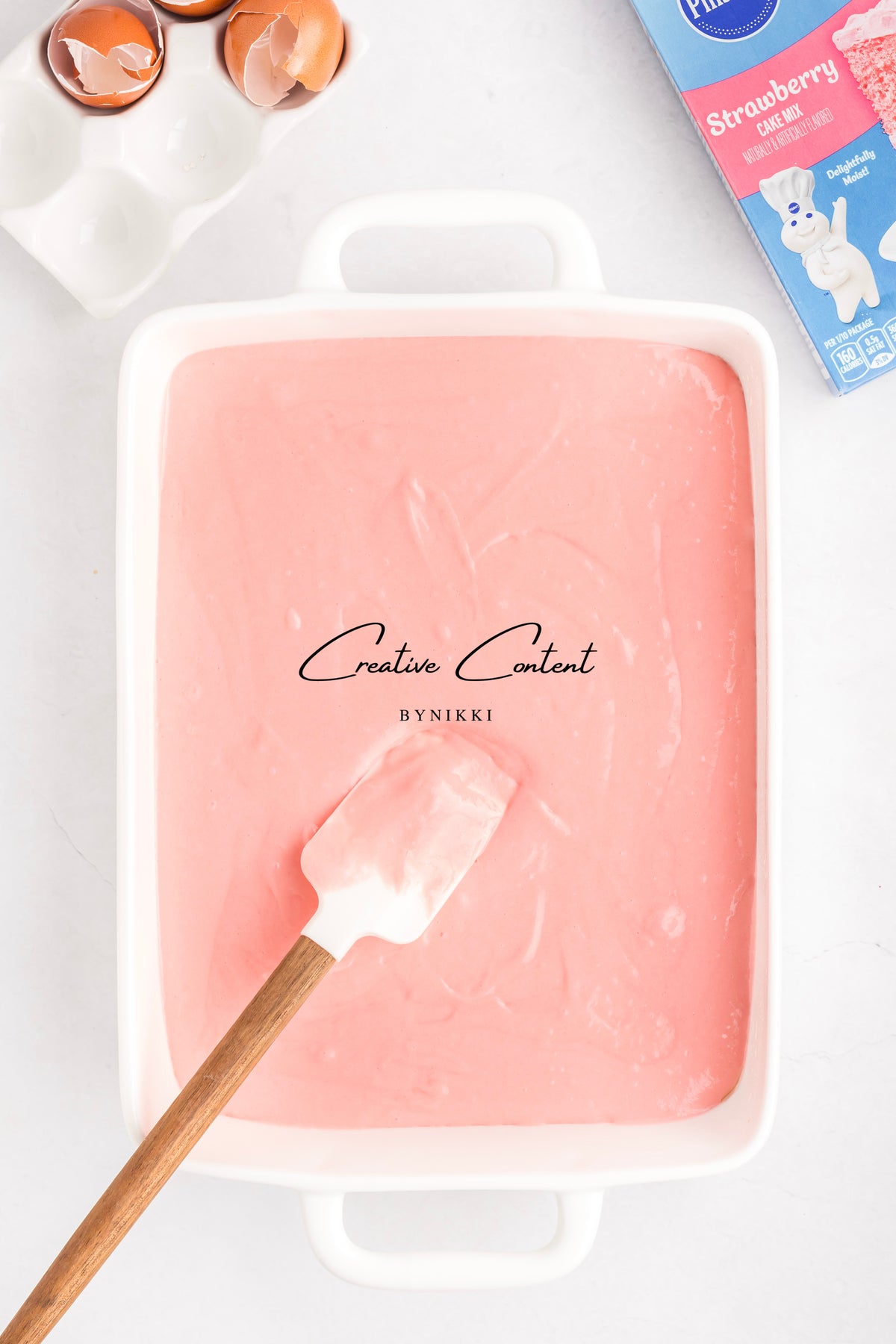 A white rectangular baking dish filled with pink batter for a Strawberry Crunch Cake - Exclusive, featuring a silicone spatula, a box of cake mix, and eggshells nearby.