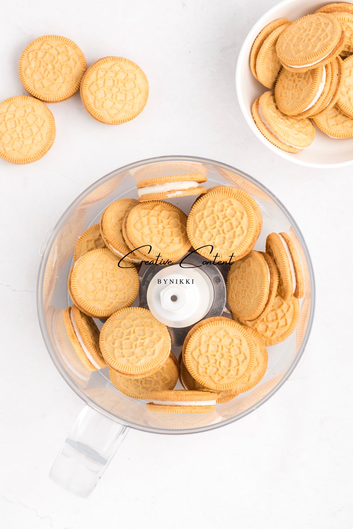 A food processor bowl filled with Strawberry Crunch Cake - Exclusive sandwich cookies, with additional cookies scattered on the surface and in a small bowl nearby.