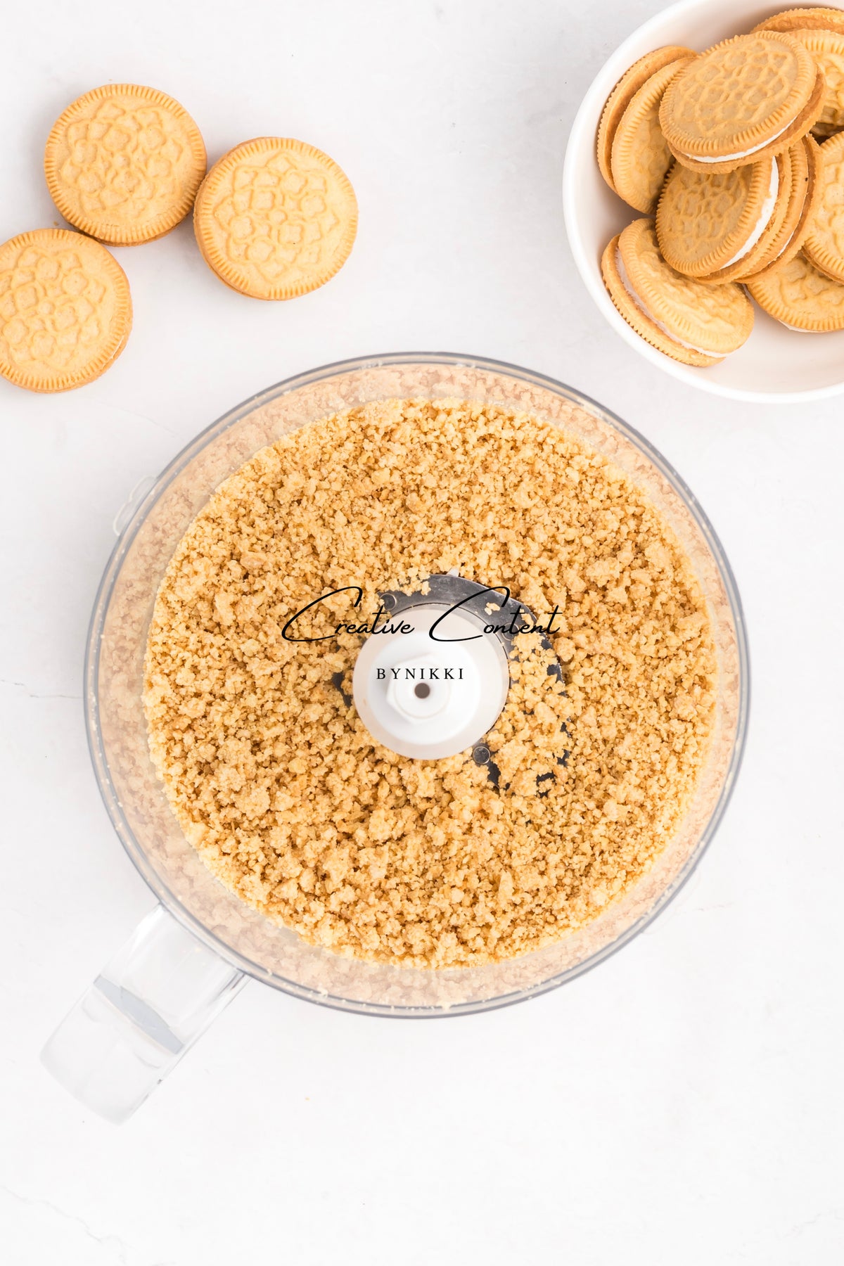 A food processor filled with crushed golden sandwich cookies used for a Strawberry Crunch Cake - Exclusive, with additional whole cookies on the side.