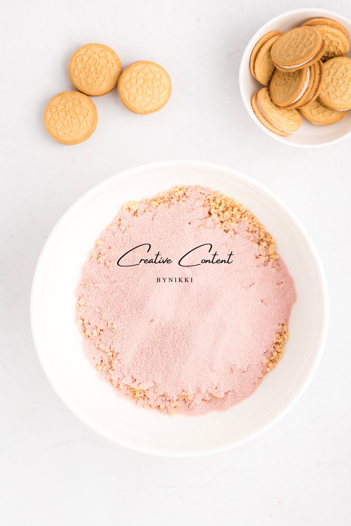 A white bowl containing Strawberry Crunch Cake - Exclusive crumbs topped with pink powder, next to a small bowl of sandwich cookies and three loose cookies on a white surface.