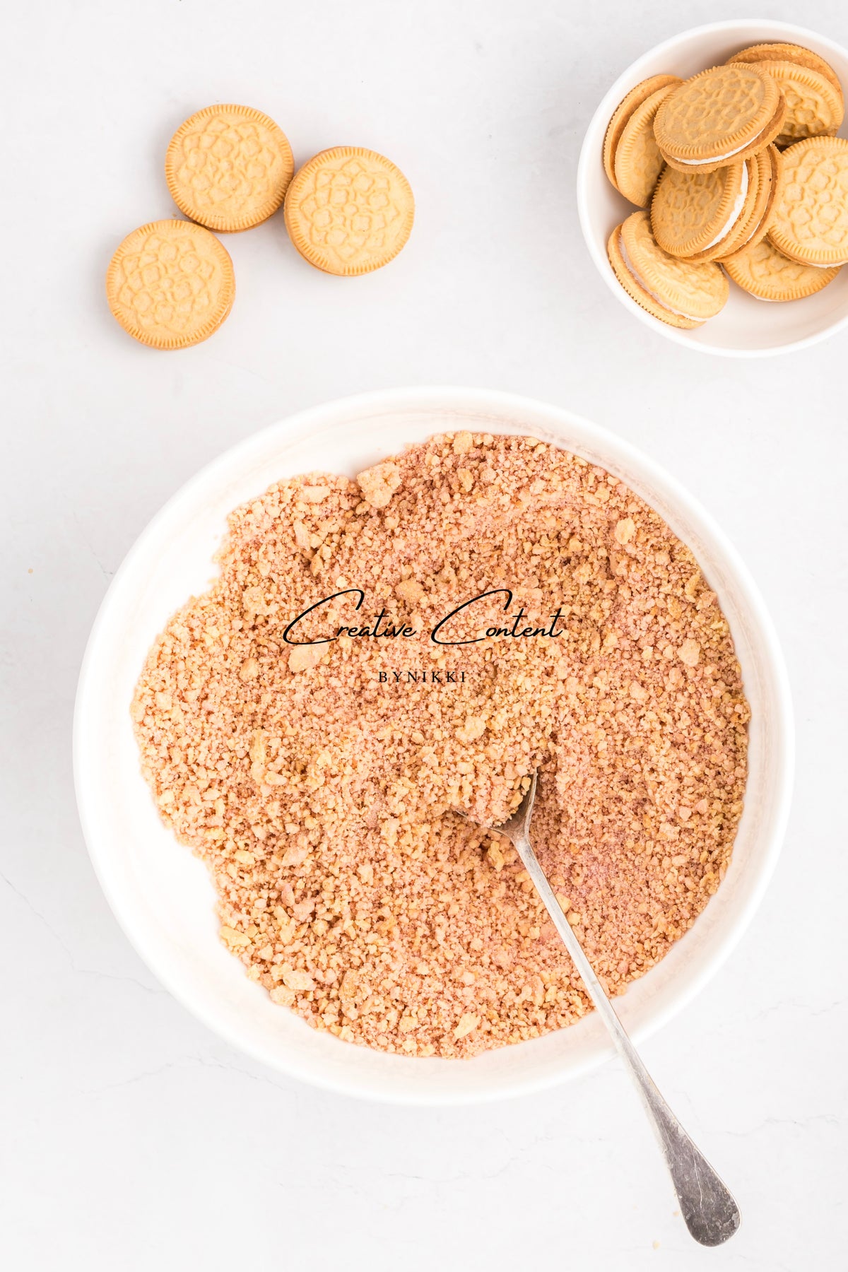 A bowl filled with Strawberry Crunch Cake - Exclusive crumbs and a spoon, accompanied by a small bowl of sandwich cookies and three loose cookies on a white surface.