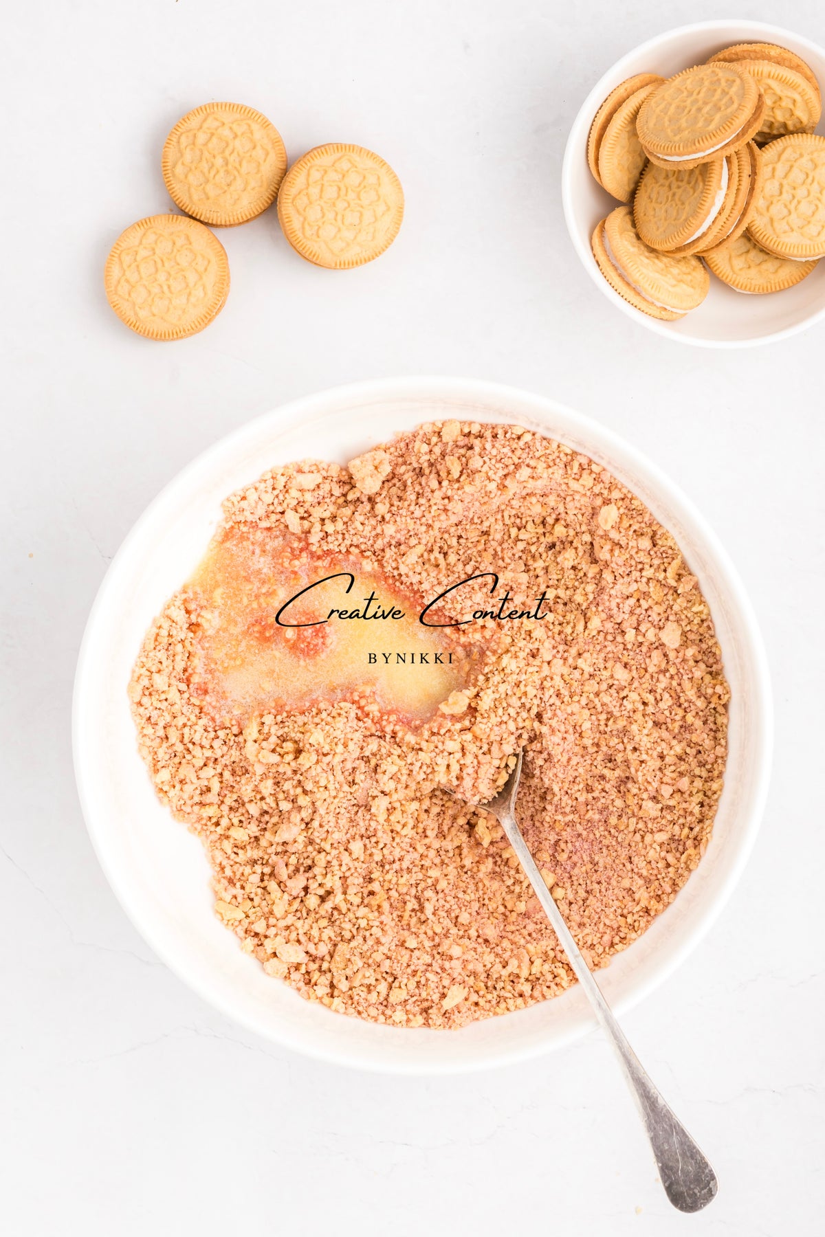 A white bowl filled with crushed cookie crumbs for a Strawberry Crunch Cake - Exclusive, with melted butter, a spoon, and whole sandwich cookies nearby.