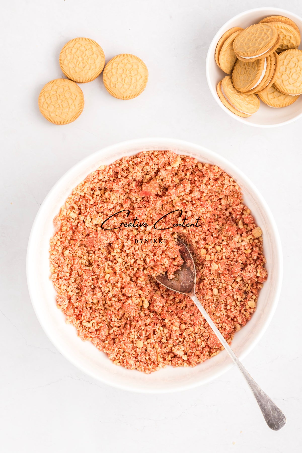 A bowl filled with Strawberry Crunch Cake - Exclusive crumbs and a metal spoon, with sandwich cookies in a small bowl and on the surface nearby.