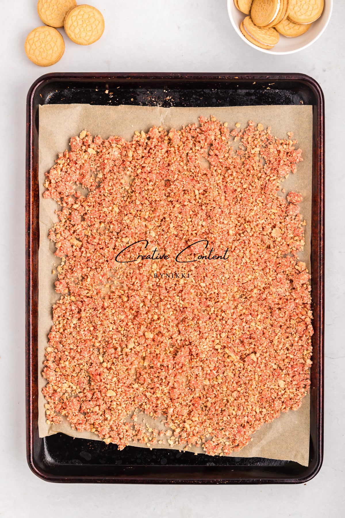 A baking sheet lined with parchment paper covered in Strawberry Crunch Cake - Exclusive crumble in pink and tan, with sandwich cookies in the corner.