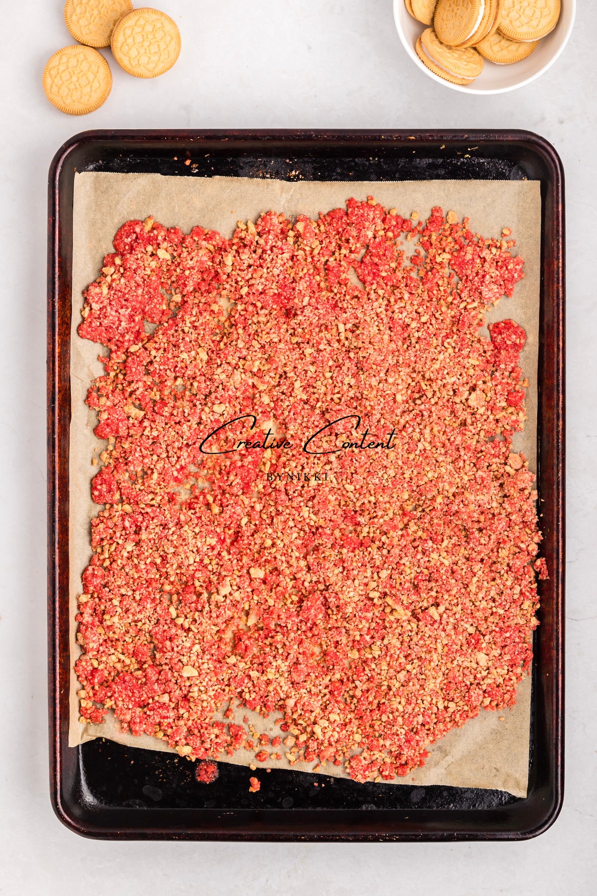 A baking sheet lined with parchment paper covered in a layer of red and tan Strawberry Crunch Cake - Exclusive crumbles, with sandwich cookies nearby.