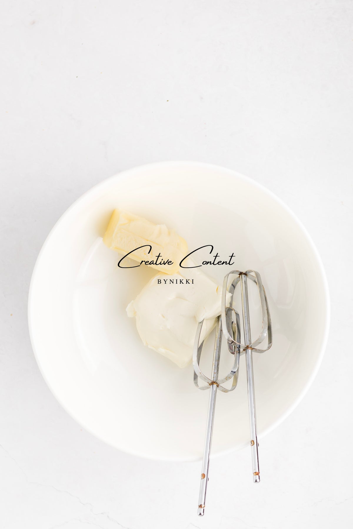 A white mixing bowl containing a block of butter and cream cheese for the Strawberry Crunch Cake - Exclusive vanilla frosting, with electric mixer beaters resting inside.