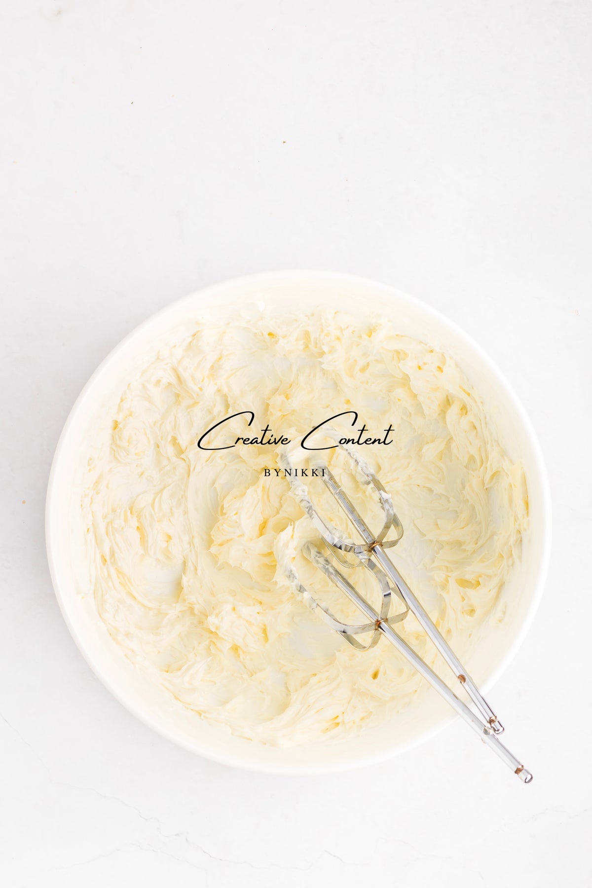 A white bowl containing pale yellow vanilla frosting for a Strawberry Crunch Cake - Exclusive, with a handheld electric mixer attachment resting inside.