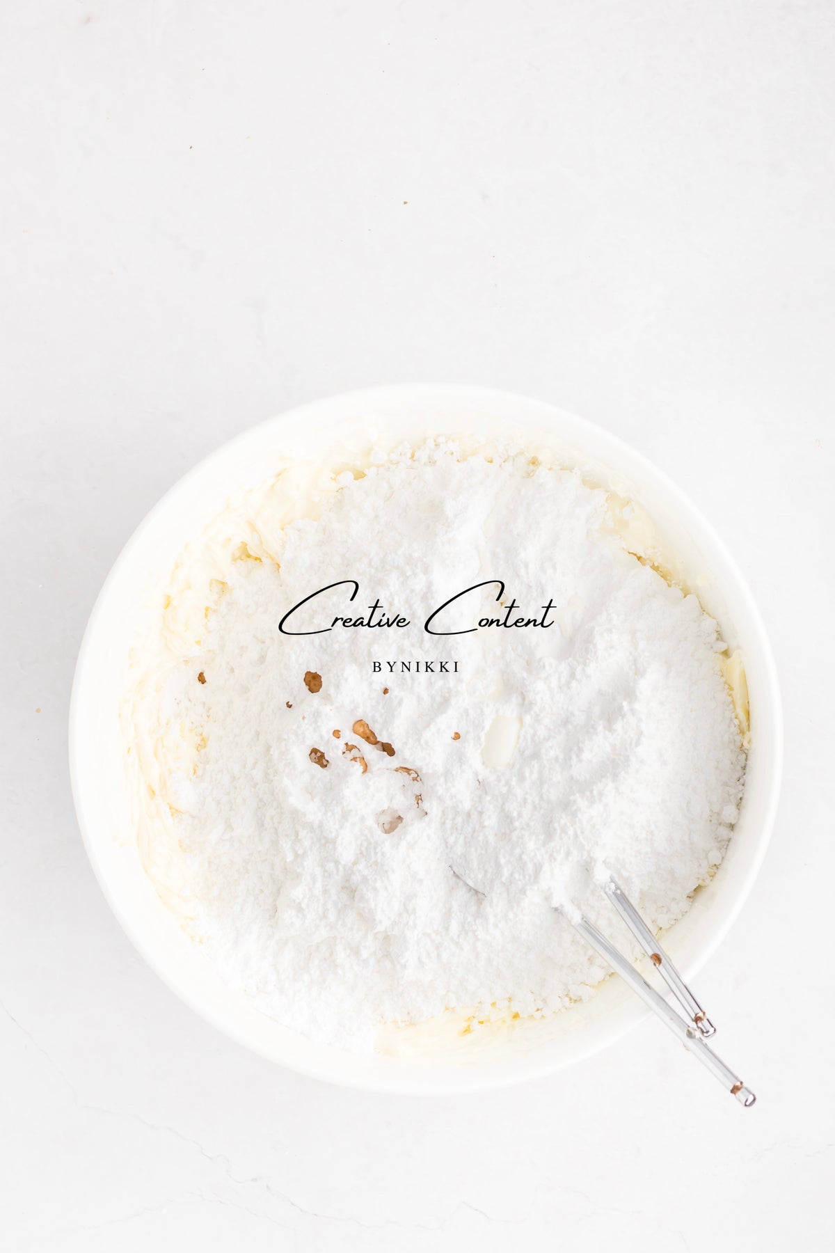 A white mixing bowl containing creamed butter and a mound of powdered sugar for Strawberry Crunch Cake - Exclusive vanilla frosting, with a metal whisk resting inside.