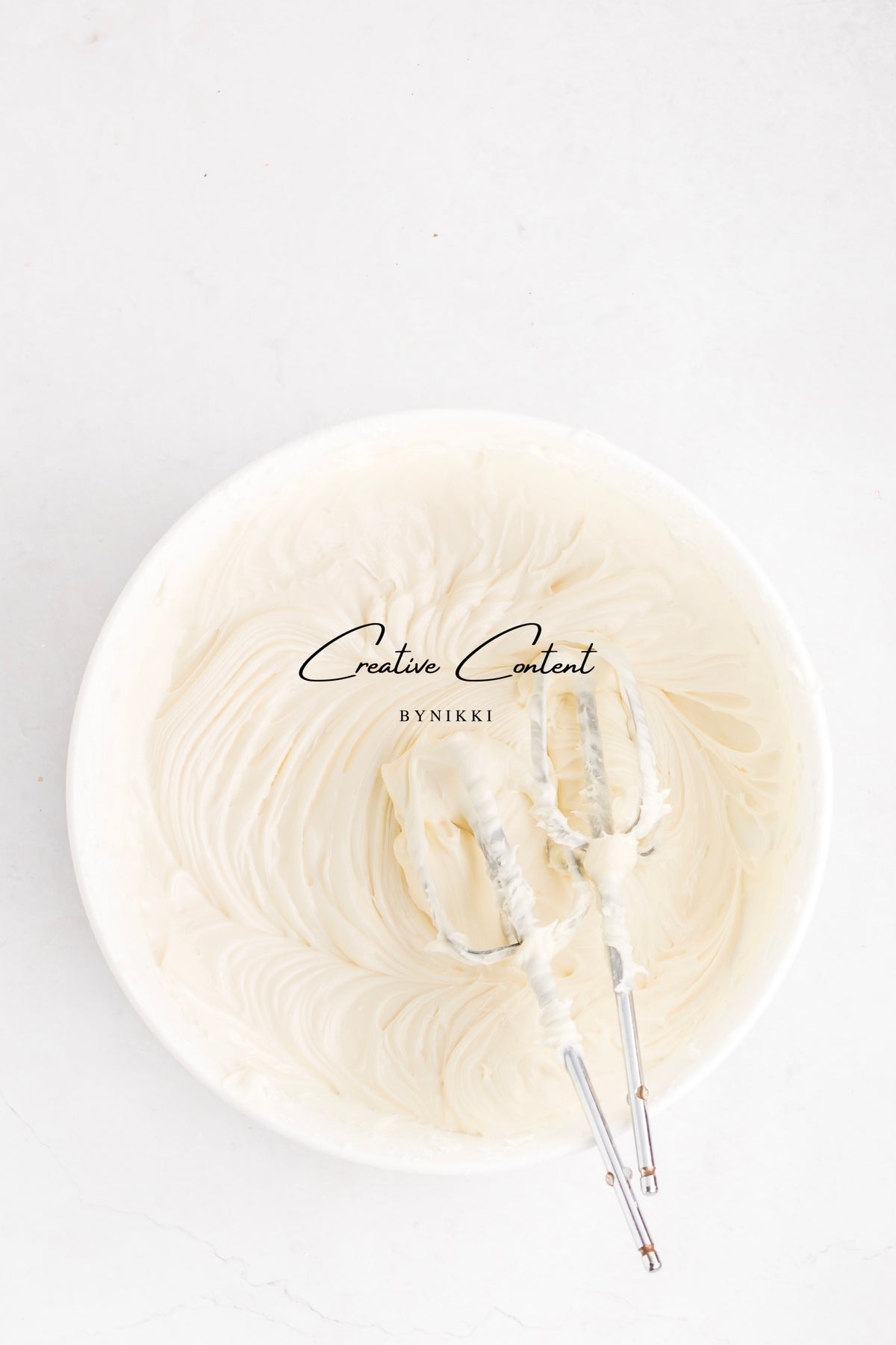 A large white bowl filled with creamy vanilla frosting and two hand mixer beaters for a Strawberry Crunch Cake - Exclusive.