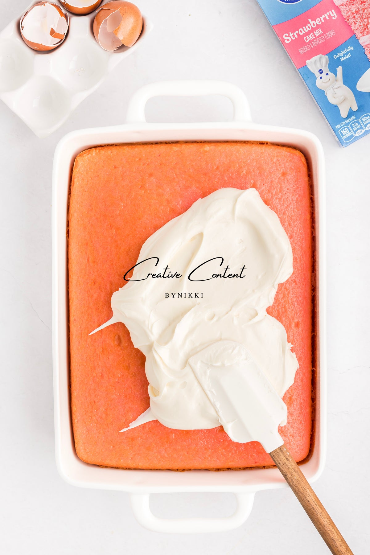A rectangular Strawberry Crunch Cake - Exclusive in a white baking dish being frosted with vanilla icing using a spatula.