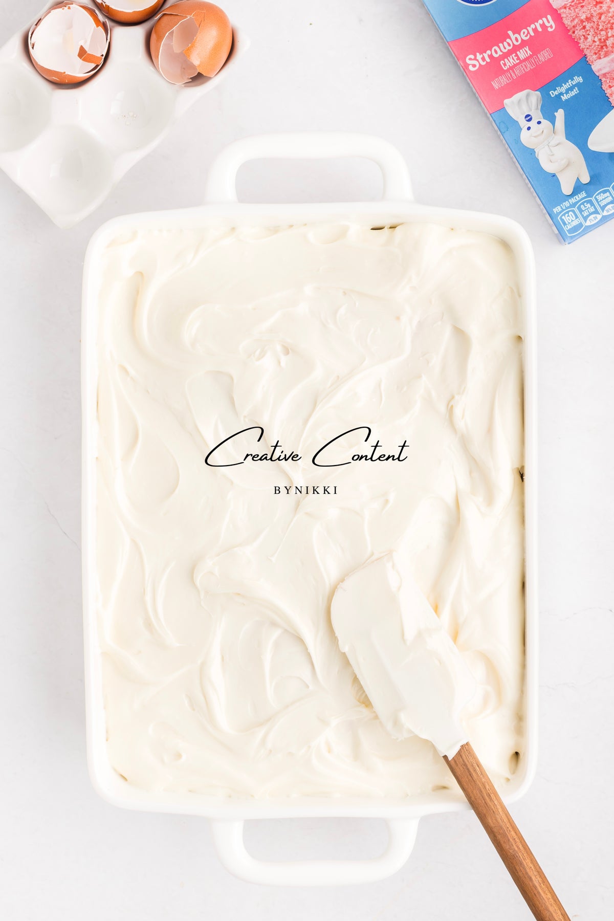 A rectangular white baking dish filled with Strawberry Crunch Cake - Exclusive topped with vanilla frosting, with a silicone spatula resting on the surface next to an egg carton and a box of strawberry cake mix.