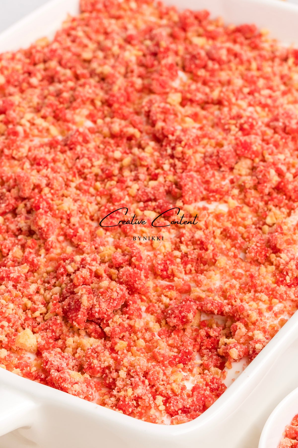 A rectangular white baking dish filled with a Strawberry Crunch Cake - Exclusive, featuring creamy layers topped with red and golden cookie crumbles.