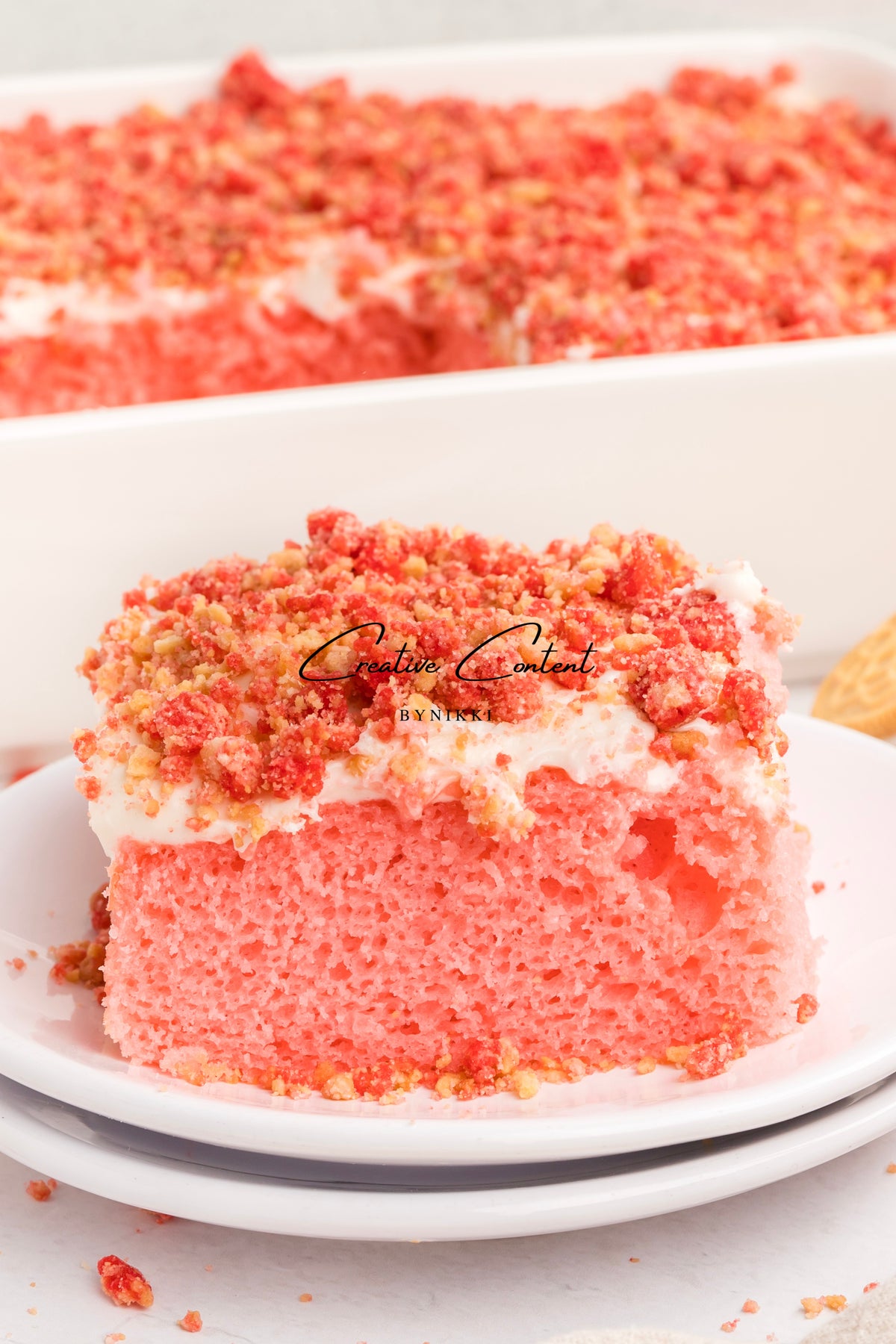A slice of Strawberry Crunch Cake - Exclusive topped with vanilla frosting and red cookie crumbles on a white plate, with a baking dish of the same cake in the background.