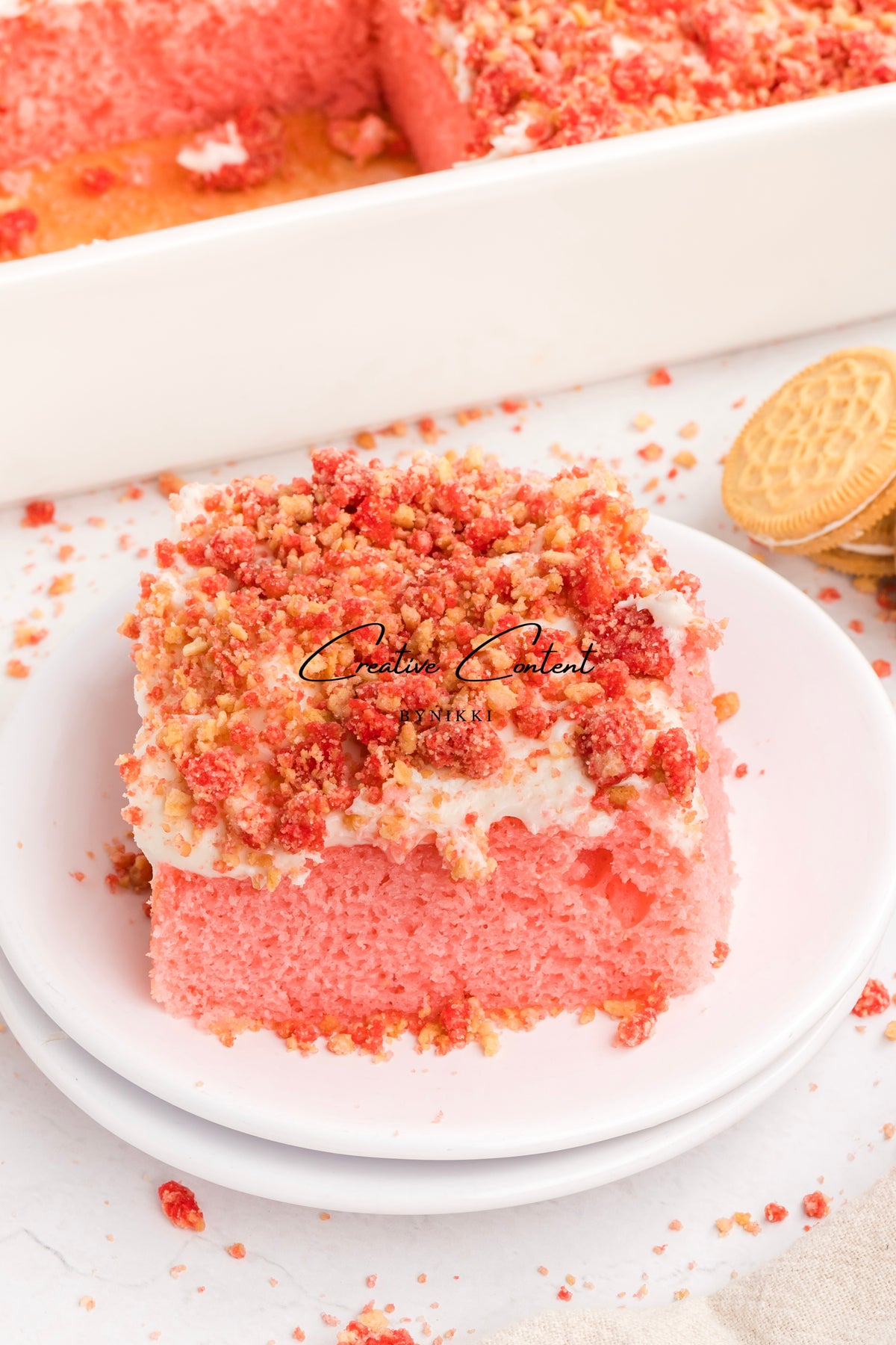 A square slice of Strawberry Crunch Cake - Exclusive topped with white frosting and red cookie crumbles on a white plate.