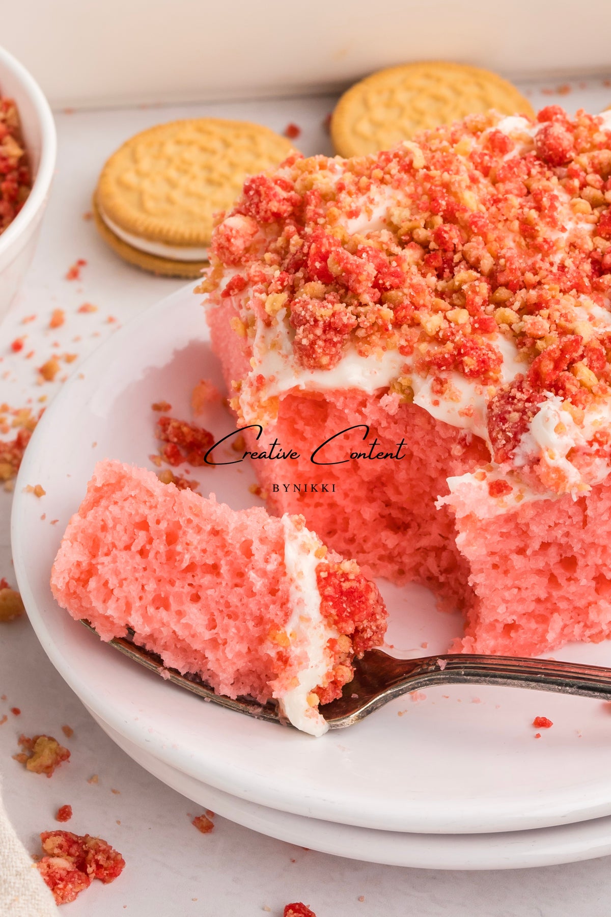 A slice of Strawberry Crunch Cake - Exclusive topped with vanilla frosting and strawberry crumbs on a silver fork, with the remaining cake and sandwich cookies on a white plate.