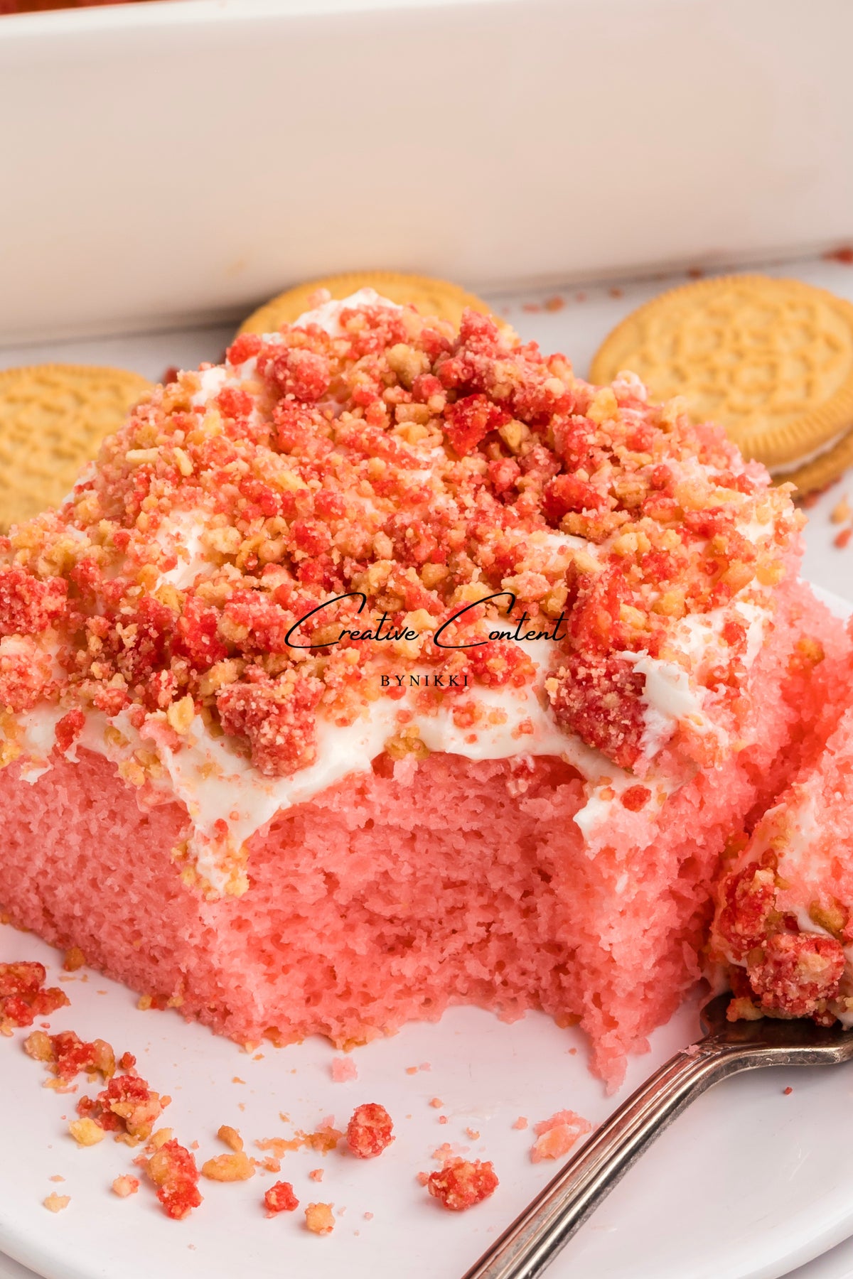 A slice of Strawberry Crunch Cake - Exclusive with bright pink cake, vanilla frosting, and red strawberry crumbles, served on a white plate with sandwich cookies.