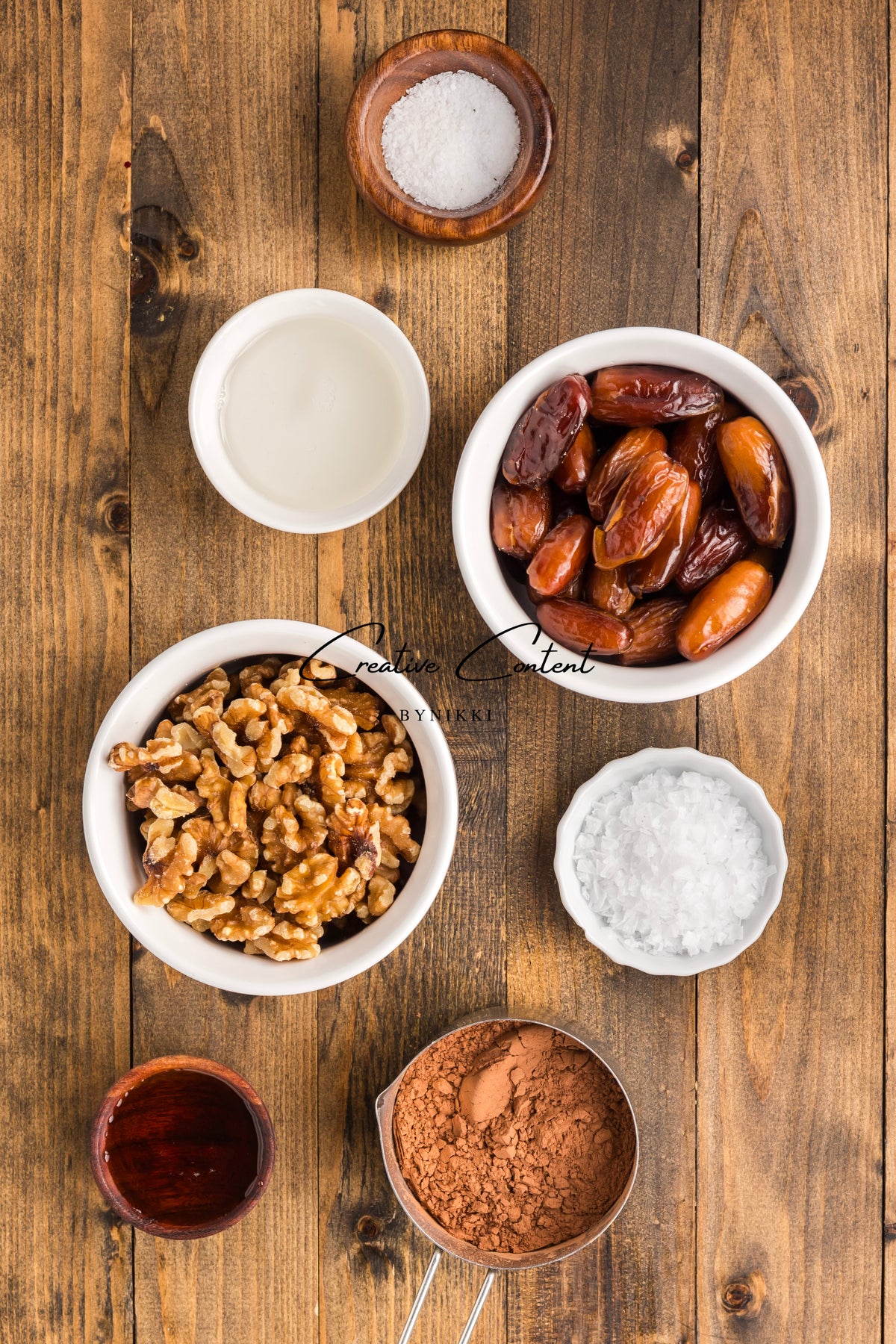Bowls containing walnuts, dates, cocoa powder, salt, and liquid ingredients for Walnut Date Bars - Exclusive arranged on a wooden surface.