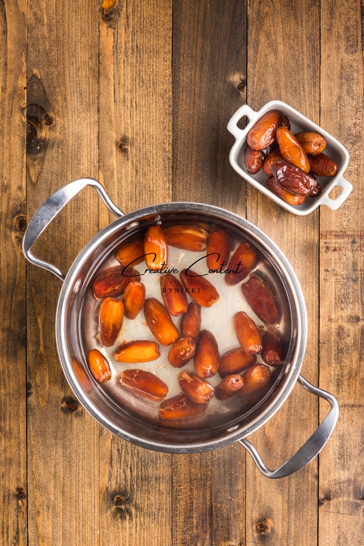 A metal pot containing dates soaked in water for Walnut Date Bars - Exclusive, with a small dish of additional dates beside it on a wooden surface.