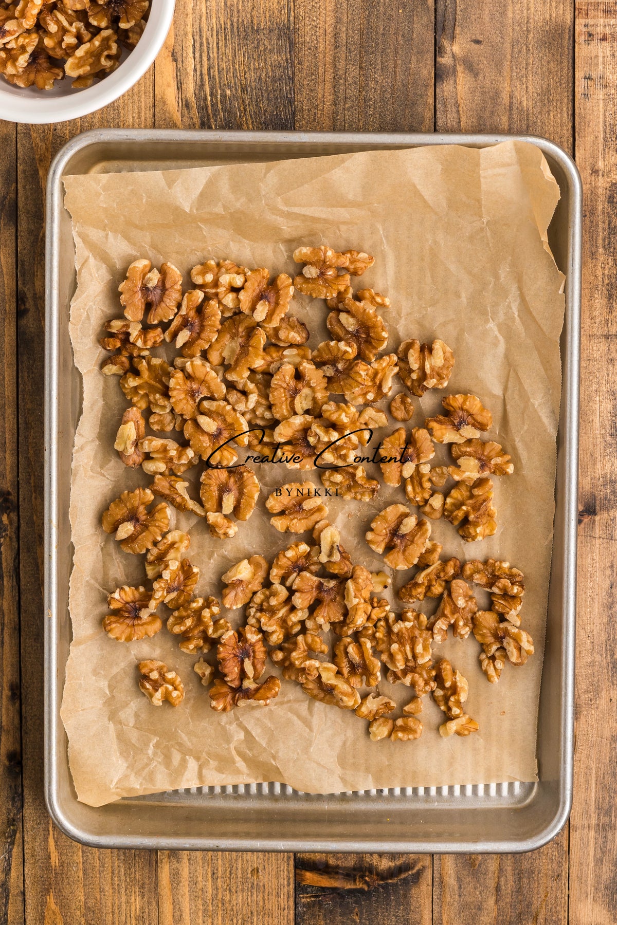 Walnut halves for Walnut Date Bars - Exclusive spread on a parchment-lined baking sheet, with a small bowl of walnuts in the corner.