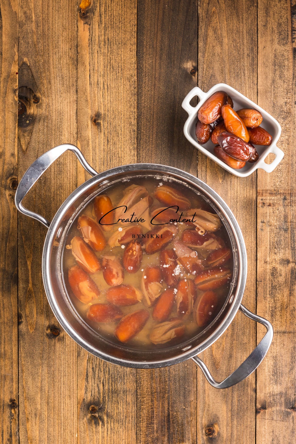 A stainless steel pot filled with dates soaking in liquid for Walnut Date Bars - Exclusive, next to a small white bowl of whole dates on a wooden surface.