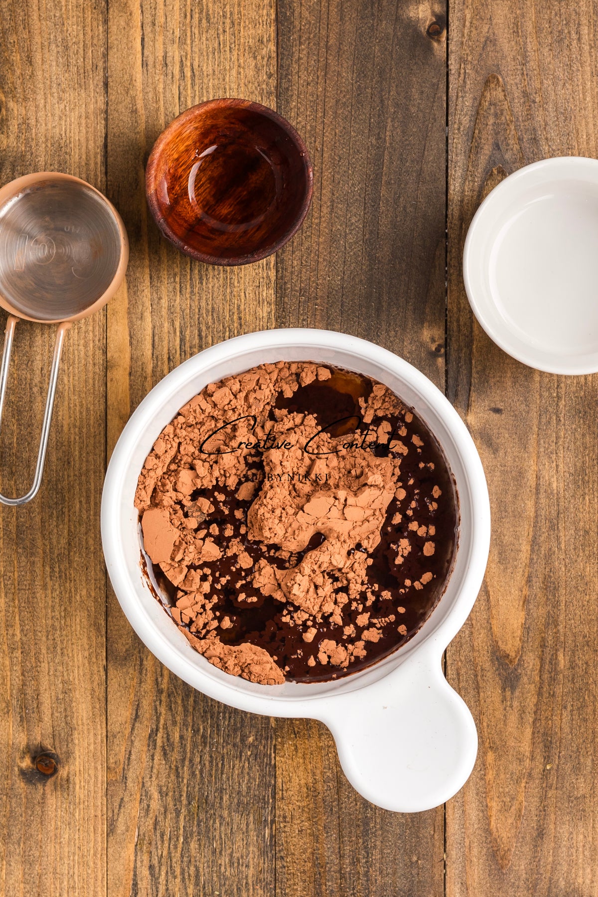 A white bowl containing dark liquid and cocoa powder for Walnut Date Bars - Exclusive, surrounded by a measuring cup, a wooden bowl, and a small white dish on a wooden surface.