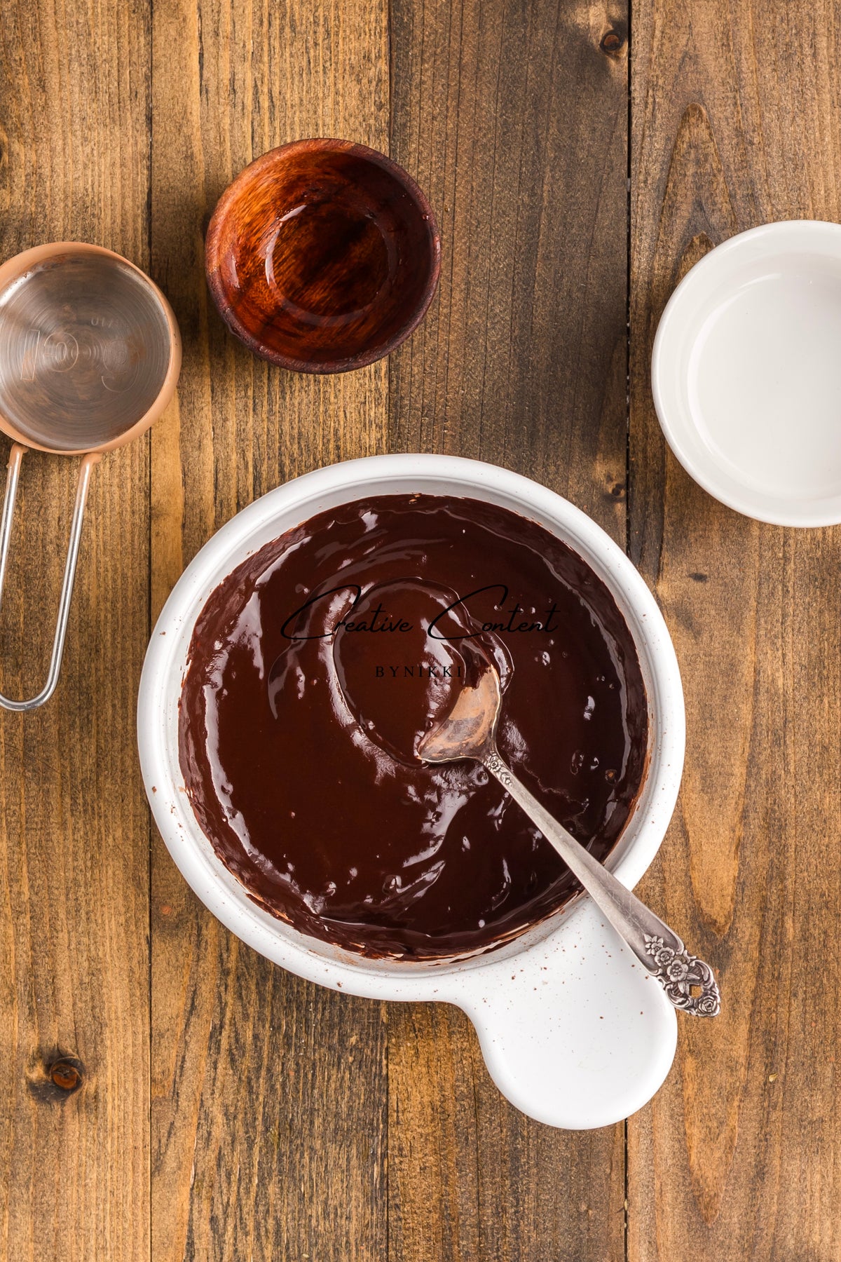 A white bowl of melted chocolate for coating Walnut Date Bars - Exclusive, with a silver spoon, a metal measuring cup, and two small bowls on a wooden surface.