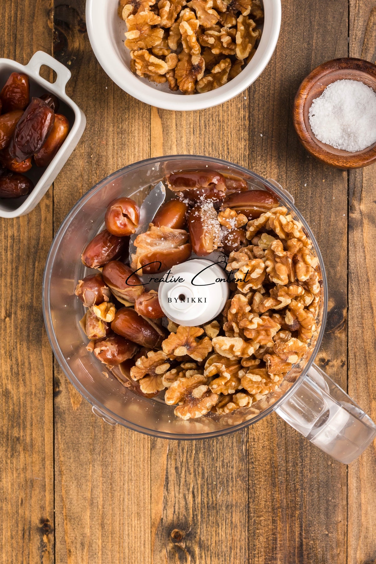 A food processor bowl filled with walnuts, dates, and a pinch of salt to make Walnut Date Bars - Exclusive, with small dishes of additional ingredients nearby on a wooden table.