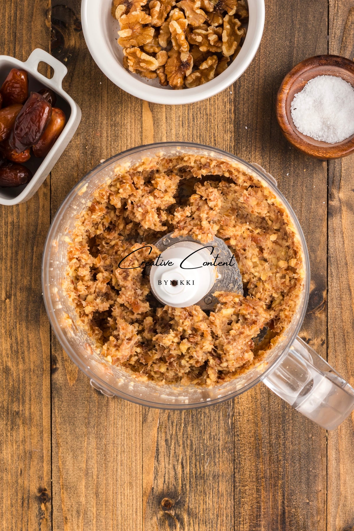 A food processor containing a mixture of chopped walnuts and dates for Walnut Date Bars - Exclusive, with bowls of whole walnuts, dates, and salt nearby.