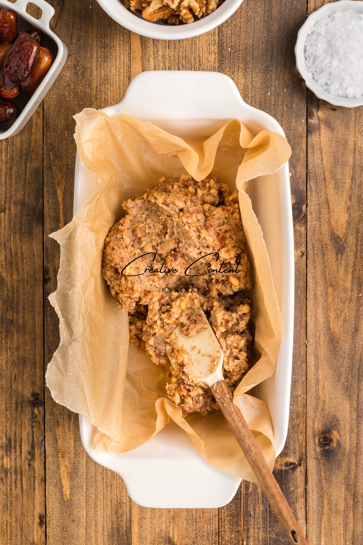 A rubber spatula presses a mixture of chopped nuts and dates into a parchment-lined baking dish to form Walnut Date Bars - Exclusive, surrounded by small bowls of ingredients.
