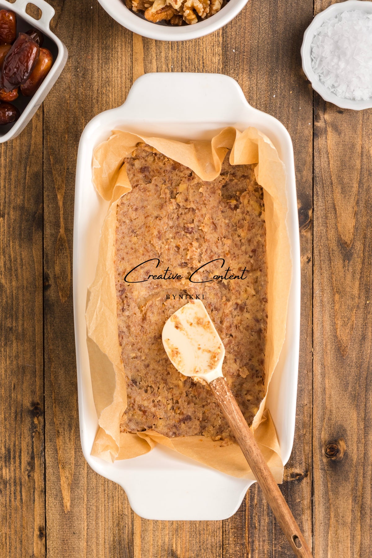 A baking dish filled with a nut and date mixture for Walnut Date Bars - Exclusive, with a silicone spatula resting on top, surrounded by bowls of dates, walnuts, and salt.