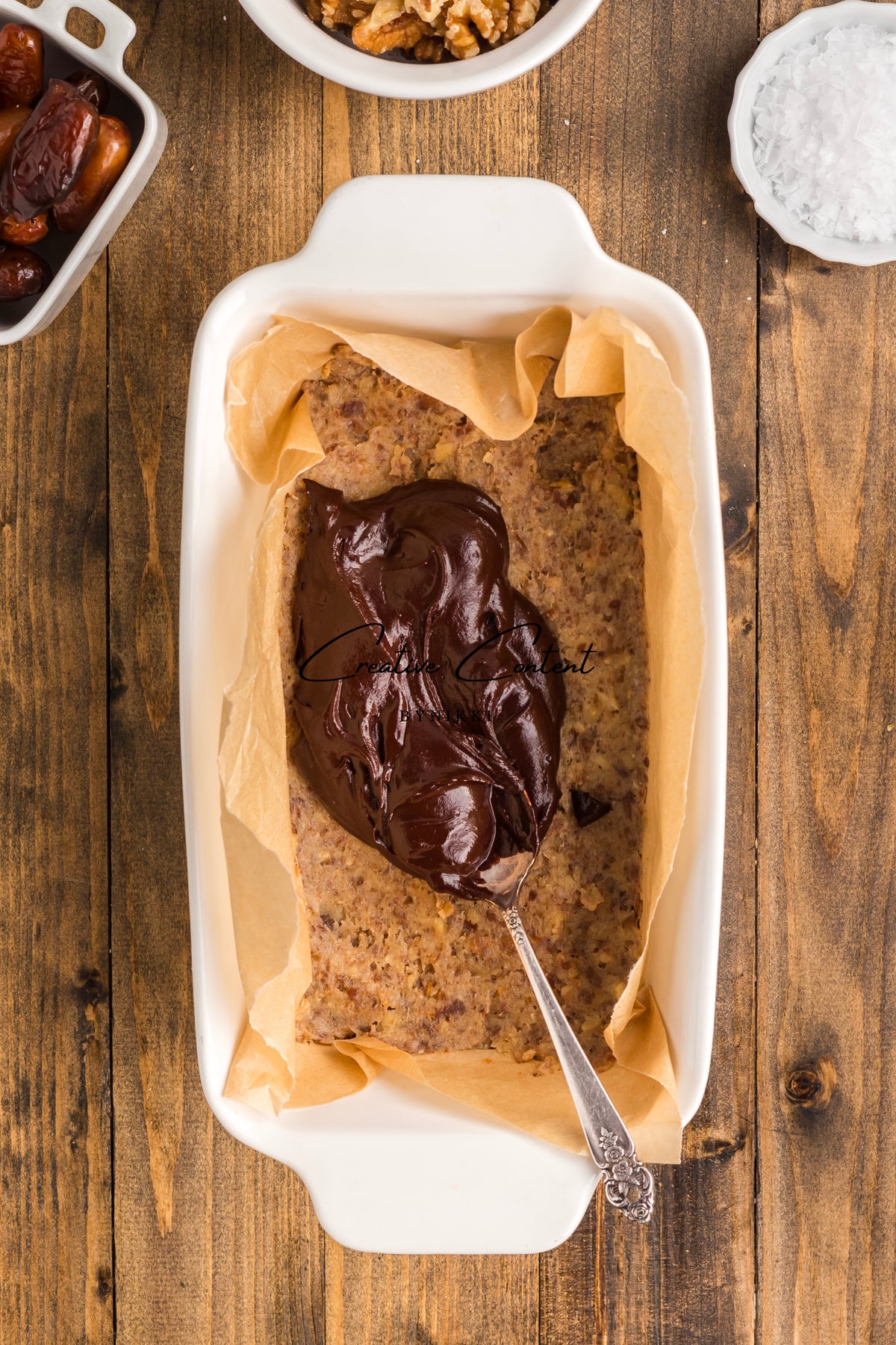 A spoon spreads chocolate ganache over a rectangular Walnut Date Bars - Exclusive in a parchment-lined baking dish, surrounded by bowls of dates, walnuts, and salt.