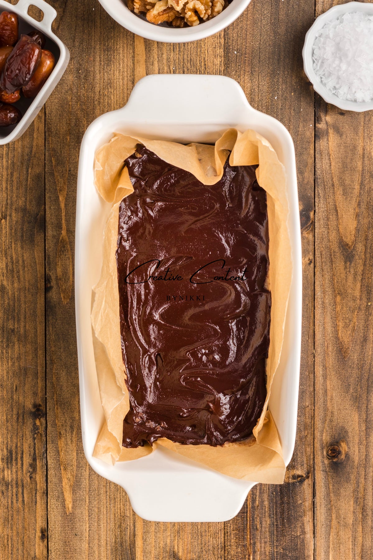A baking dish lined with parchment paper filled with chocolate batter for Walnut Date Bars - Exclusive, surrounded by small bowls of dates, walnuts, and sea salt.