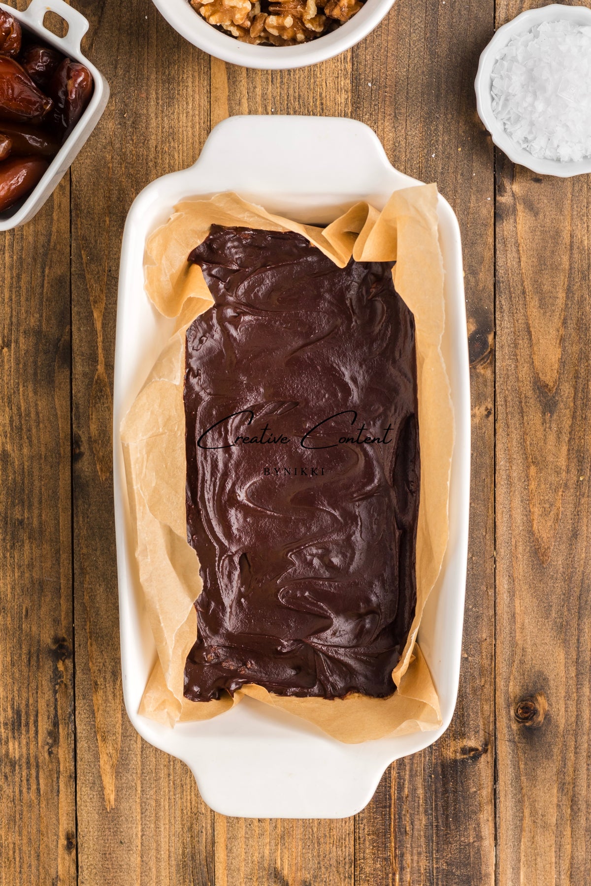 A rectangular baking dish lined with parchment paper containing Walnut Date Bars - Exclusive, surrounded by small bowls of walnuts, dates, and sea salt.
