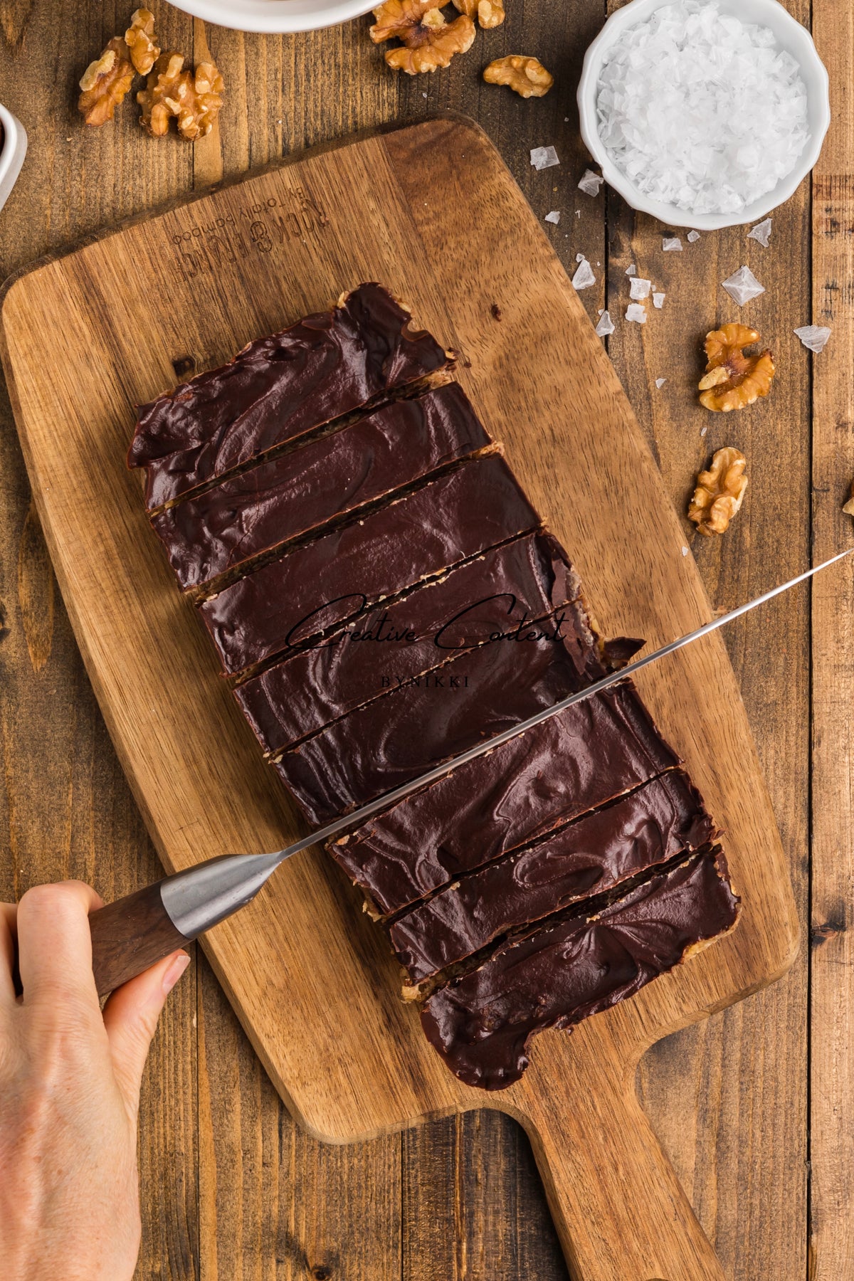 A hand uses a knife to slice a rectangular Walnut Date Bars - Exclusive on a wooden cutting board, surrounded by scattered walnuts and salt.