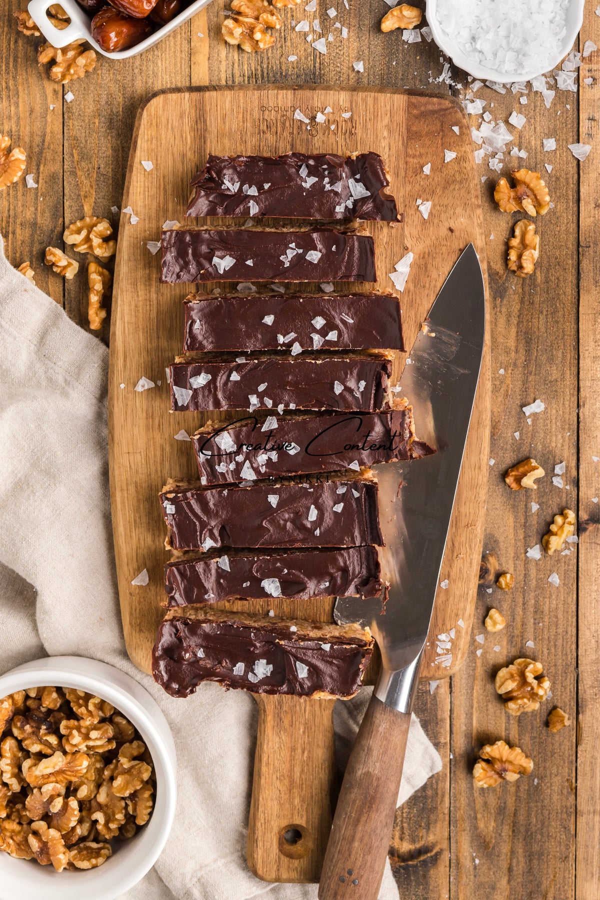 Sliced Walnut Date Bars - Exclusive coated in chocolate and sprinkled with sea salt on a wooden cutting board with a chef’s knife.
