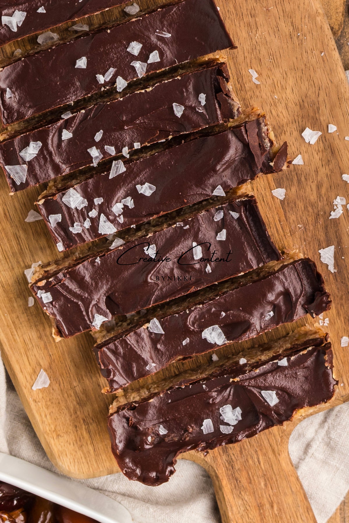 Sliced Walnut Date Bars - Exclusive coated in chocolate and topped with dark chocolate ganache and sea salt flakes on a wooden board.