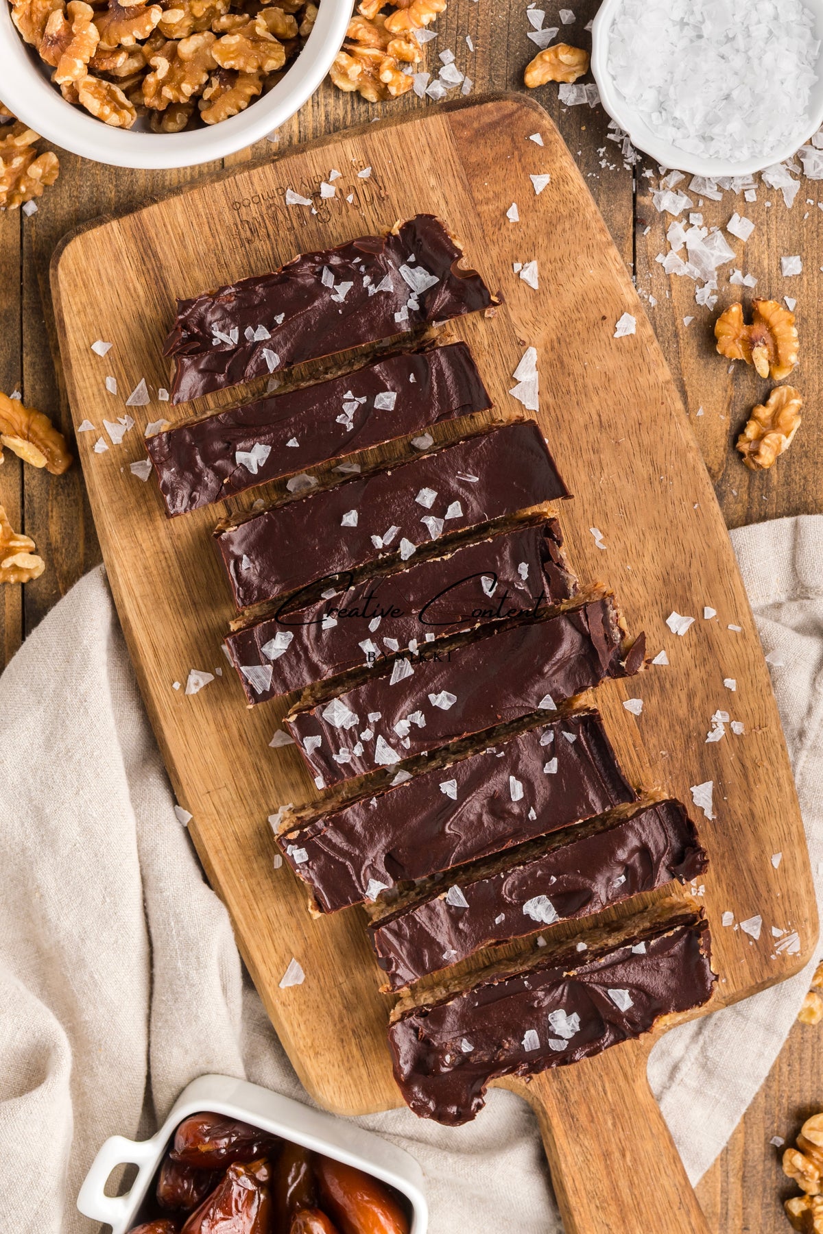 Six Walnut Date Bars - Exclusive coated in chocolate and sprinkled with sea salt on a wooden cutting board, surrounded by bowls of walnuts and dates.