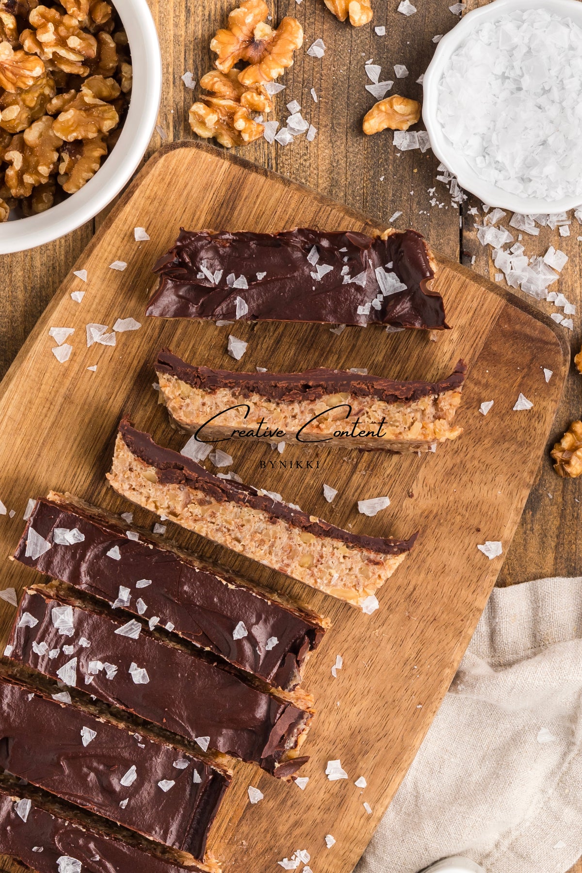 Sliced Walnut Date Bars - Exclusive coated in chocolate and sprinkled with sea salt on a wooden cutting board, with bowls of walnuts and salt nearby.