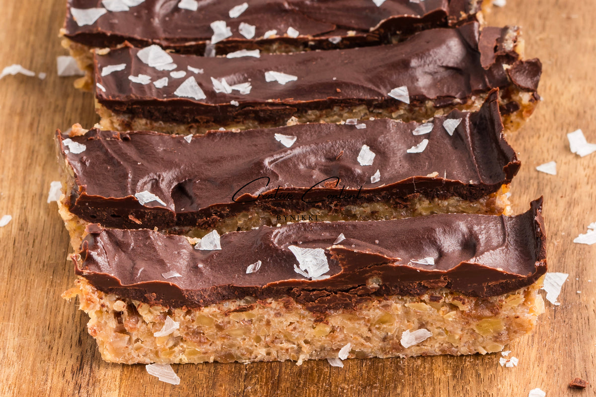 Four rectangular Walnut Date Bars - Exclusive topped with dark chocolate and sea salt flakes, arranged on a wooden board.