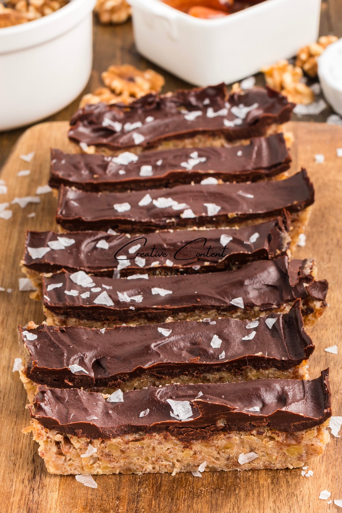 Walnut Date Bars - Exclusive: rectangular chocolate coated bars with a nutty base, topped with frosting and flaky sea salt, arranged on a wooden cutting board.