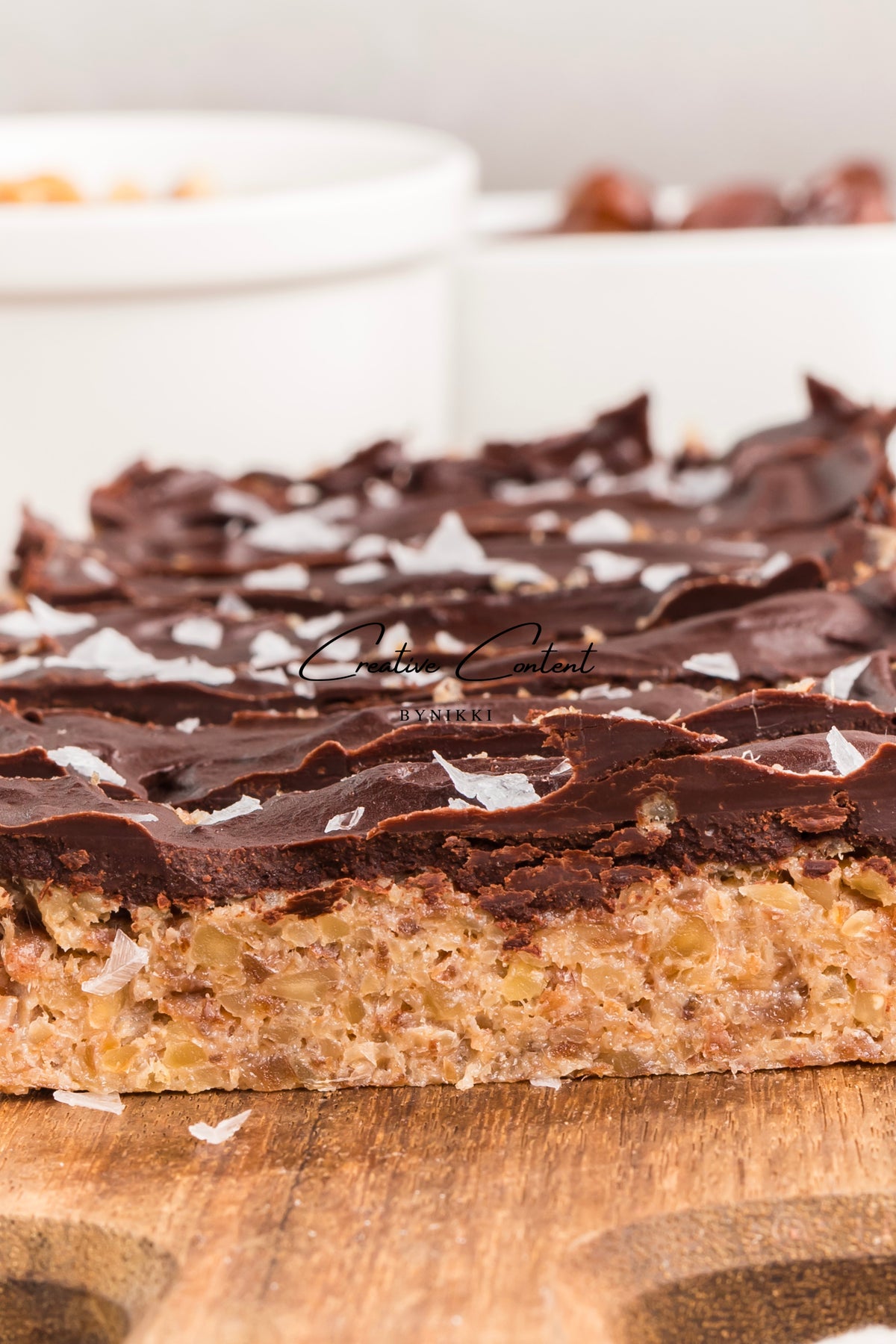 A rectangular Walnut Date Bars - Exclusive with a dense, textured base topped with a layer of dark chocolate and flaky sea salt.