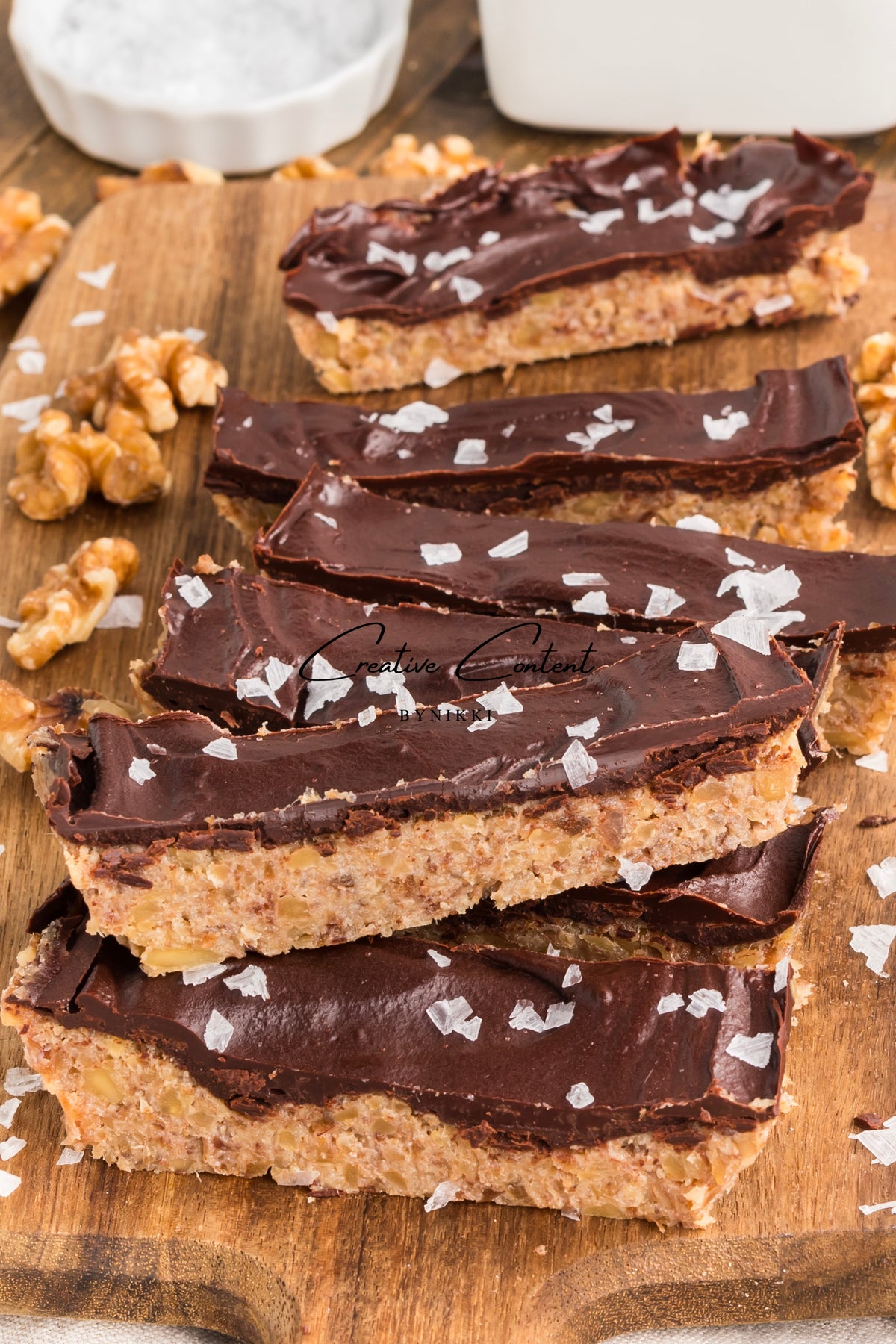 A stack of Walnut Date Bars - Exclusive topped with dark chocolate ganache and sea salt on a wooden board with scattered walnut pieces.