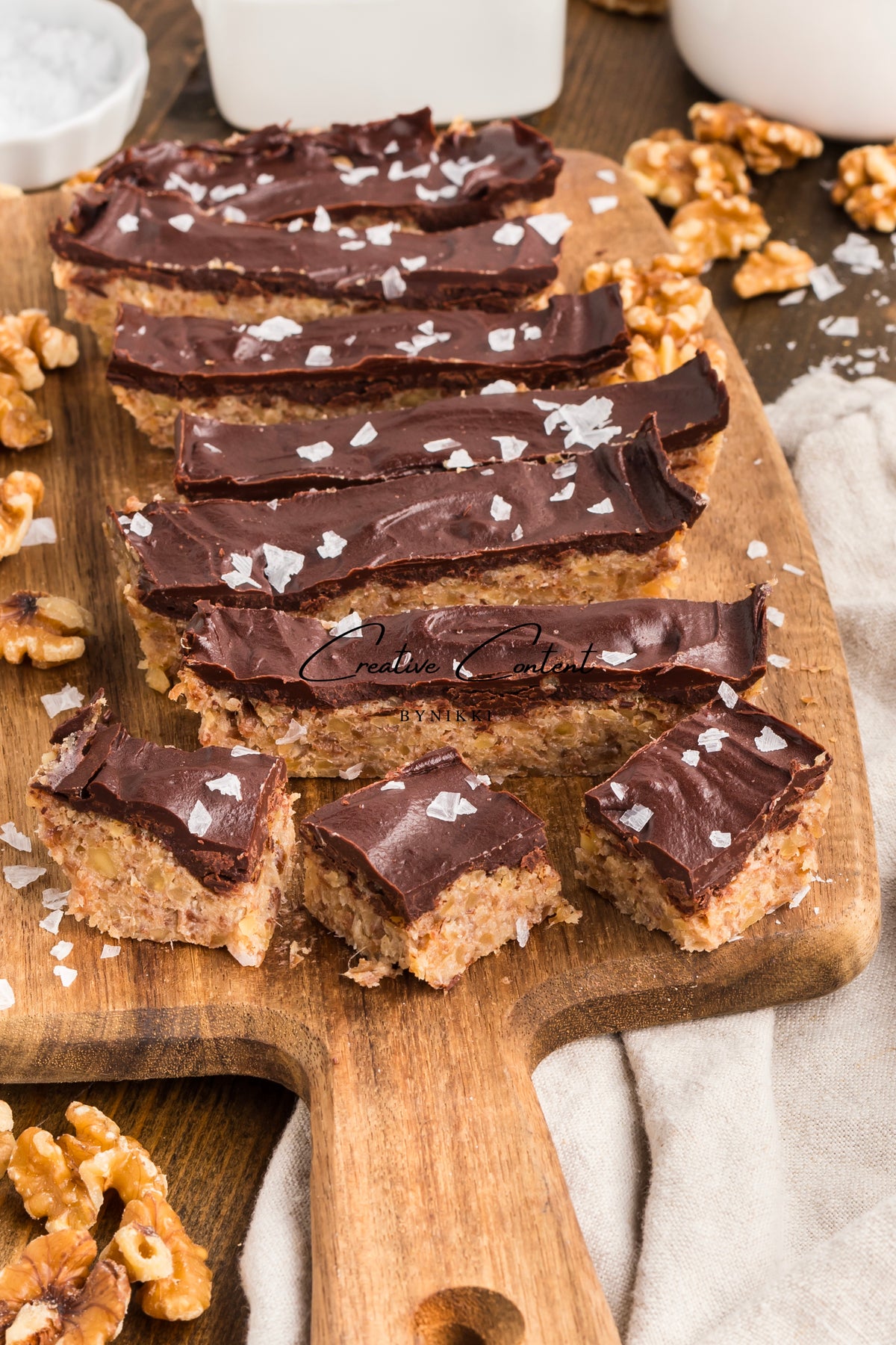 Walnut Date Bars - Exclusive: naturally sweetened date and walnut bars coated in dark chocolate and sprinkled with sea salt, arranged on a wooden cutting board with loose walnut pieces.